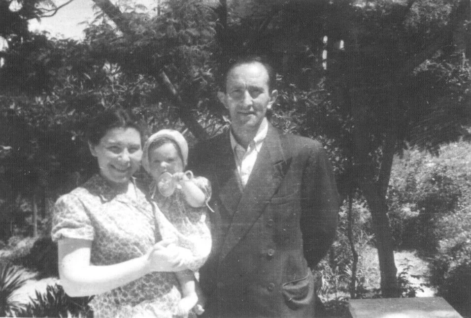 Masha holding young Shifra, with Aryeh standing beside them outdoors.