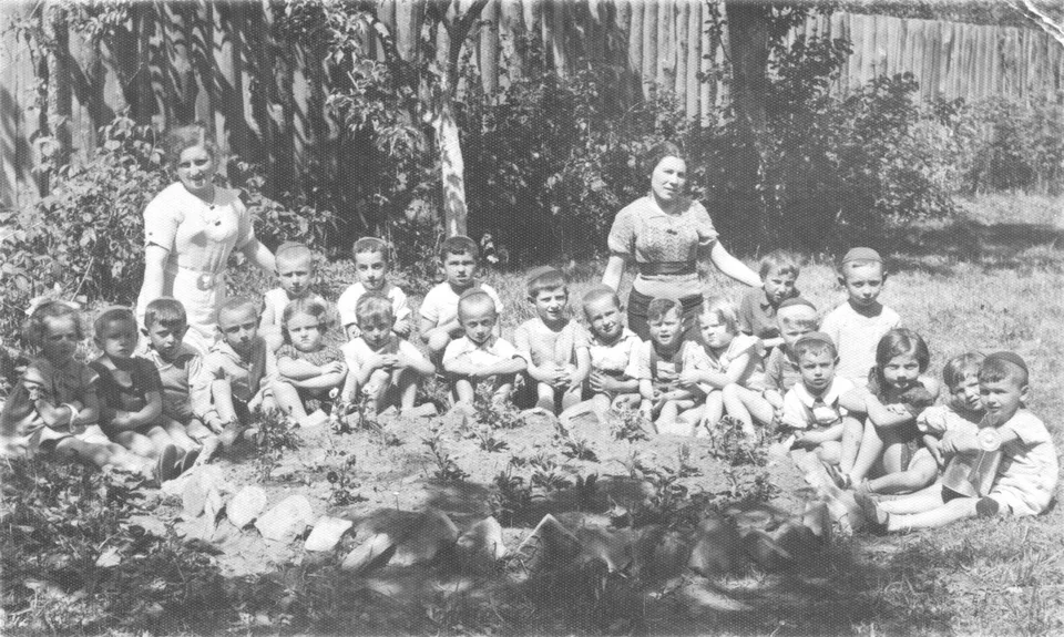 A group of children and two women photographed outdoors beside a wooden building.