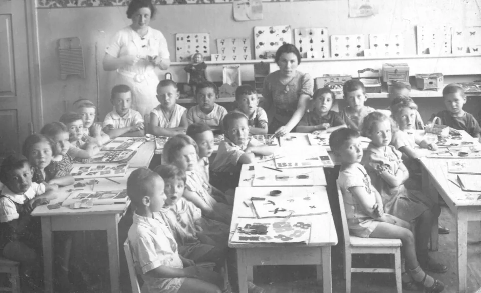 Children seated at classroom tables with two women beside them.