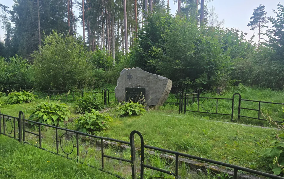 The memorial stone within a fenced grassy site surrounded by trees.